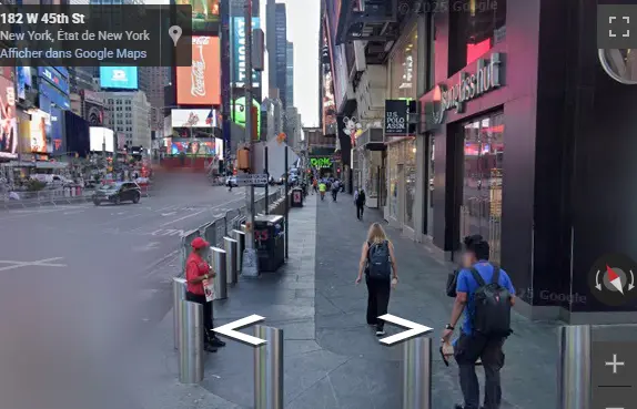 Rue animée de Times Square, New York (182 W 45th St) capturée via Google Street View, mélangeant effervescence urbaine et détails réalistes. Au premier plan, des bornes métalliques délimitent le trottoir où des piétons (dont une femme avec sac à dos) circulent devant des enseignes lumineuses emblématiques (publicités géantes, néons). À gauche, un agent en gilet rouge surveille la zone, ajoutant une touche d’authenticité new-yorkaise. En arrière-plan, gratte-ciels illuminés, taxis jaunes et écrans géants rappellent l’énergie 24/7 du quartier. Mots-clés : Times Square vue rue, ambiance New York City, Google Maps Street View, vie urbaine Manhattan, enseignes lumineuses NYC, tourisme virtuel États-Unis.