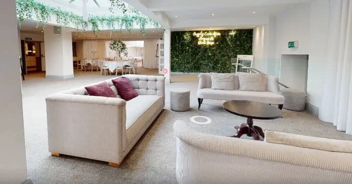 *&#34;Salle de réception élégante et lumineuse, alliant design contemporain et touches naturelles pour un espace polyvalent idéal pour événements, mariages ou réunions professionnelles. L’image met en valeur un salon d’accueil spacieux avec deux canapés en tissu beige assortis de coussins bordeaux, disposés autour d’une table basse ronde en métal et marbre. En arrière-plan, un mur végétal verdoyant apporte une ambiance apaisante et moderne, tandis que des lustres suspendus et une verrière inondent l’espace de lumière naturelle. À gauche, une salle à manger élégante avec des tables rondes nappées de blanc et des chaises design est visible, parfaite pour des dîners ou des conférences. Les sol moquetté beige et les murs blancs épurés renforcent le côté luxe discret et intemporel de ce lieu. Mots-clés associés : salle de réception mariage, espace événementiel lumineux, salon d’accueil contemporain, mur végétal intérieur, décoration élégante pour événements, lieu polyvalent mariage/séminaire, ambiance luxe et nature.&#34;*