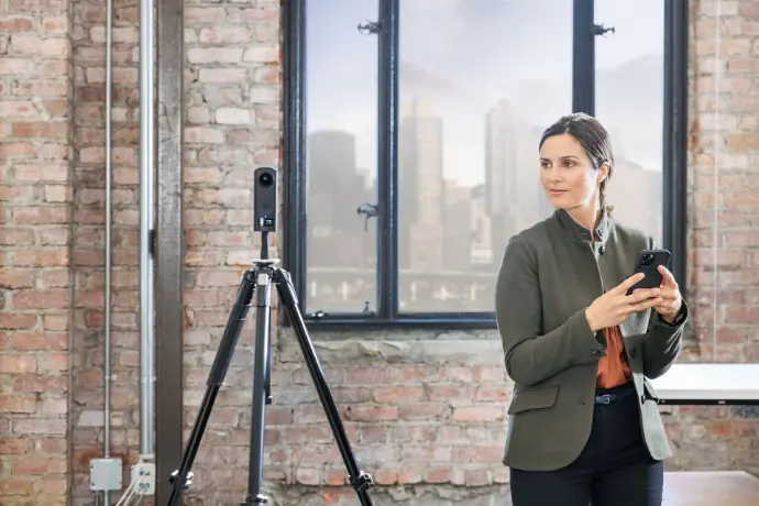 Une professionnelle en tenue élégante, vêtue d&#39;une veste verte, utilise un smartphone tout en se tenant près d&#39;un trépied équipé d&#39;une caméra ou d&#39;un capteur. L&#39;environnement, avec ses murs en briques apparentes et ses grandes fenêtres, évoque un espace de travail moderne et créatif. Cette scène illustre parfaitement les domaines de la réalité virtuelle, de la captation 3D, des présentations interactives, ou encore des visites virtuelles. Idéal pour représenter les innovations technologiques dans les secteurs de l&#39;immobilier, de l&#39;événementiel ou du marketing digital.