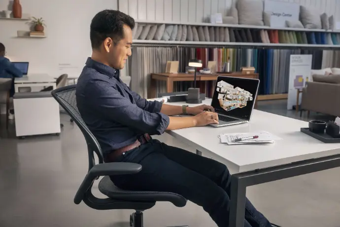 Un professionnel assis à un bureau moderne, travaillant sur un ordinateur portable affichant un plan 3D d’aménagement intérieur. Vêtu d’une chemise bleue, il semble concentré sur la conception ou la modification d’un projet de design d’espace, probablement pour un salon ou un espace commercial. Autour de lui, un environnement de travail bien organisé avec des échantillons de tissus et des éléments de décoration en arrière-plan, évoquant un bureau de design d’intérieur ou un showroom. Cette scène illustre parfaitement les métiers de l’architecture d’intérieur, du design spatial, et de la planification de projets créatifs pour les professionnels du secteur.