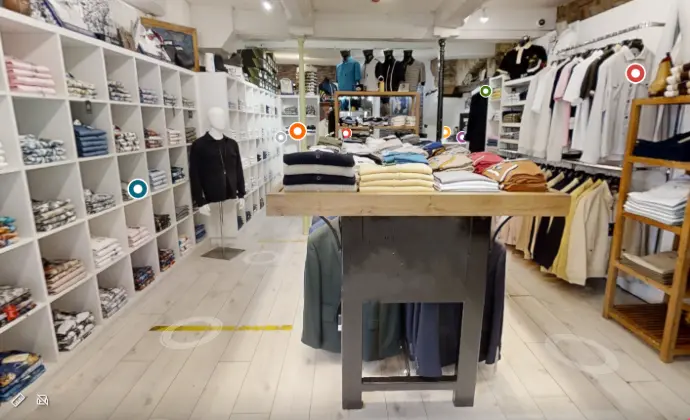 Intérieur d’un magasin de vêtements moderne et organisé, spécialisé dans la vente de prêt-à-porter masculin et féminin. L’espace lumineux et épuré met en valeur des étagères remplies de vêtements soigneusement pliés, tels que des t-shirts, pulls, chemises et accessoires. Au centre, une table d’exposition présente des piles de pulls et sweats colorés, tandis que des mannequins habillés illustrent les tendances actuelles. À droite, des porte-manteaux en bois affichent des vestes et manteaux élégants, et des rayons muraux proposent des vêtements triés par taille et par couleur. Le sol en bois clair et les murs blancs renforcent l’ambiance chaleureuse et accueillante du magasin, idéale pour une expérience shopping agréable.