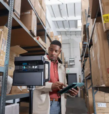 Un employé en train d’effectuer un inventaire dans un entrepôt logistique moderne. Vêtu d’une veste claire et équipé d’un terminal portable ou d’un scanner, il vérifie les colis et les produits stockés sur des étagères métalliques remplies de cartons. L’entrepôt, organisé et bien éclairé, illustre une gestion efficace des stocks et des processus logistiques. Cette scène est représentative des métiers de la logistique, de la gestion des stocks, et de l’optimisation des entrepôts, essentiels pour les entreprises de commerce et de distribution.