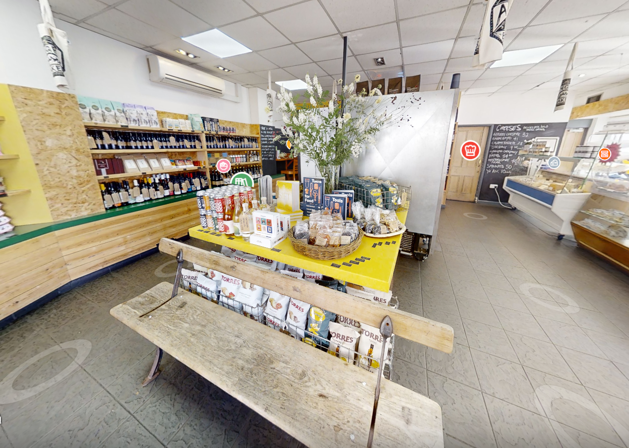 Intérieur chaleureux et accueillant d’une épicerie fine et boutique gourmande, spécialisée dans la vente de produits locaux, vins, et spécialités artisanales. L’espace lumineux et bien organisé met en valeur des étagères en bois remplies de bouteilles de vin, d’huiles d’olive, de condiments et de conserves. Au centre, une table jaune vif expose des paniers garnis de biscuits, confiseries, et produits secs, tandis qu’un comptoir frigorifique présente une sélection de fromages et charcuteries. À gauche, un large choix de boissons et d’accessoires culinaires est soigneusement disposé, et à droite, une vitrine réfrigérée met en avant des plats préparés et desserts frais. Une ardoise murale affiche les promotions et suggestions du jour, ajoutant une touche conviviale à cette ambiance d’épicerie de quartier authentique et moderne.