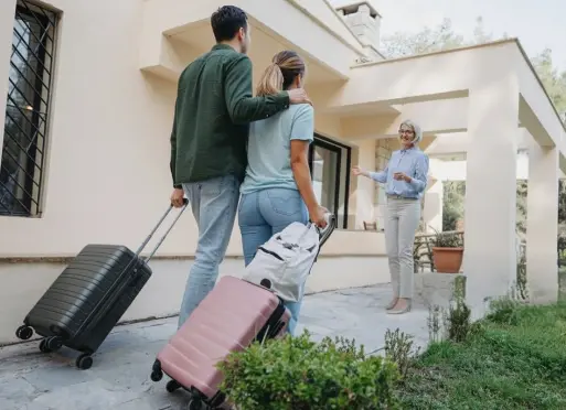 Couple de voyageurs arrivant dans une villa de vacances moderne avec valises, accueillis par la propriétaire souriante. Scène d&#39;arrivée dans un hébergement de luxe avec architecture contemporaine et jardin paysager.